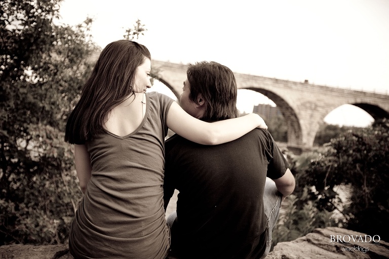 engaged couple sitting arm in arm in front of stone arch bridge in sepia photograph